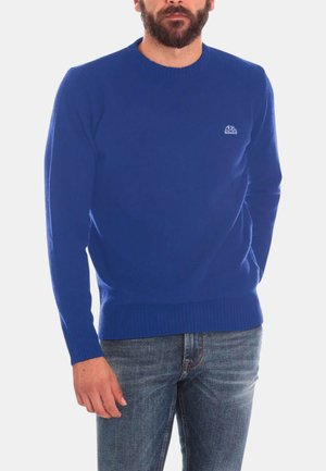 Uomo con barba che indossa un maglione blu a maniche lunghe e jeans blu, in piedi con una mano parzialmente nella tasca.