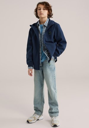 Fiú áll zsebre tett kézzel, tengerészkék dzsekit, kék kockás inget, kék pólót, világos farmert és fehér tornacipőt visel.