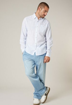 Homme debout les mains dans les poches, portant une chemise à rayures bleues claires à boutons, un pantalon bleu et des baskets blanches avec des rayures noires.