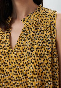 Žluté plisované šaty s výstřihem do V, zdobené černobílým abstraktním vzorem. Lehká látka, texturované záhyby a detail s volánkovým límečkem.