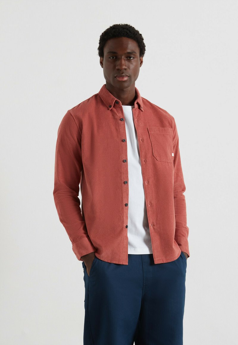 Camicia button-up in corduroy rosso con una singola tasca sul petto, abbinata a una t-shirt bianca e pantaloni blu navy, con bottoni neri e tessuto strutturato.