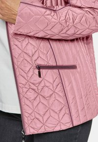 Gequilted roze jas met een diagonaal patroon, een ritszak aan de zijkant en contrasterende afwerking, gemaakt van een zachte, lichte stof.