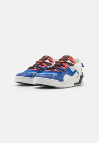 Blå och vita träningsskor har en strukturerad ovandel, röda detaljer, svarta snören och en kraftig sula med en distinkt svart bakre hälpanel.