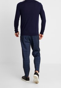 Man som går bort klädd i en marinblå tröja, mörkblå rutiga byxor och svarta sneakers med vita sulor, på vit bakgrund.