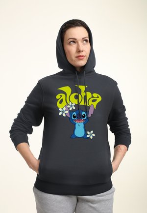 Jonge vrouw die een donkere hoodie draagt met cartoonfiguur Stitch en de tekst "aloha", handen in de zakken, kijkt omhoog naar de zijkant.