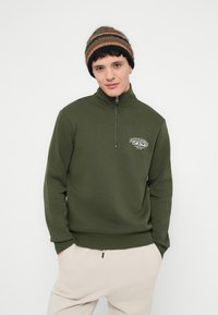 Φούτερ με φερμουάρ σε ελιά πράσινο χρώμα με ψηλό γιακά, με λευκό λογότυπο στο στήθος. Συνδυάζεται με ανοιχτό μπεζ παντελόνι, το μοντέλο φοράει ριγέ σκουφάκι.