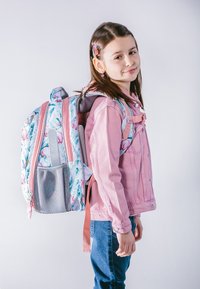 Plecak w kwiatowy wzór, w jasnoróżowych i szarych kolorach, z wyściełanymi ramiączkami oraz siateczkową kieszenią; noszony na plecach dziecka w różowej kurtce dżinsowej.