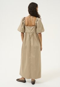 Robe maxi en beige clair, avec des manches bouffantes, un dos nu avec des liens, et une taille froncée. Portée avec des ballerines en mesh noires.