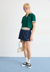 Polo cropped vert avec des rayures blanches, jupe plissée marine, baskets noir et blanc, et lunettes de soleil miroir roses, tenant un petit sac crème.