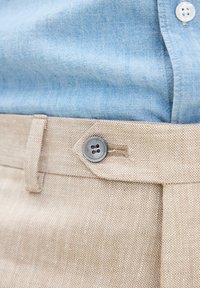 Beige strukturierte Hose mit einem am Bund befestigten Knopf, kombiniert mit einem hellblauen Hemd, das in die Hose gesteckt ist.