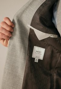 Helle graue Karoblazer mit einem glatten, braunen Futter, das ein dezentes geometrisches Muster aufweist. Enthält ein Etikett mit der Aufschrift "NEXT TAILORING."