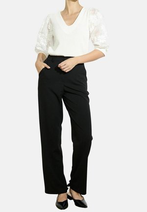 Kobieta ubrana w biały fakturowany top z bufkami z koronki, czarne szerokie spodnie z wysokim stanem oraz czarne spiczaste buty typu slingback.