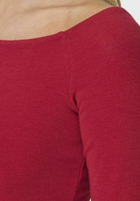 Crvena rebrasta majica dugih rukava s širokim izrezom, s mekim teksturom i prilagođenim siluetom. Vidljive su šavne detalje na ramenima.