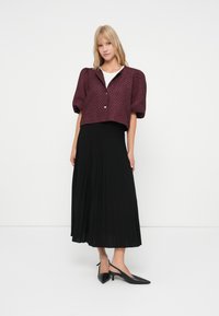 Frau in einer strukturierten pflaumenfarbenen Kurzarmjacke, weißem Oberteil, langem schwarzen plissierten Rock und schwarzen spitzen Slingback-Pumps vor weißem Hintergrund.