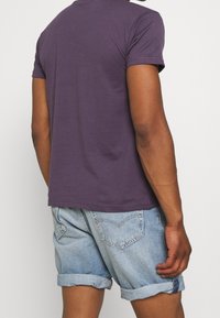 Fialové bavlněné tričko s krátkým rukávem, zobrazené z zadní strany, ve spojení se světle modrými denimovými šortkami s ohrnutými nohavicemi a zadními kapsami.