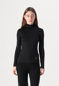 Chandail à col roulé noir à manches longues en tissu doux, avec une coupe ajustée et un logo triangulaire discret dans le coin inférieur droit.