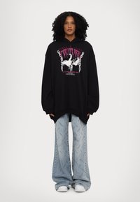 Black oversized hoodie with graphic design featuring white and pink text. Paired with light blue wide-leg jeans displaying branding.