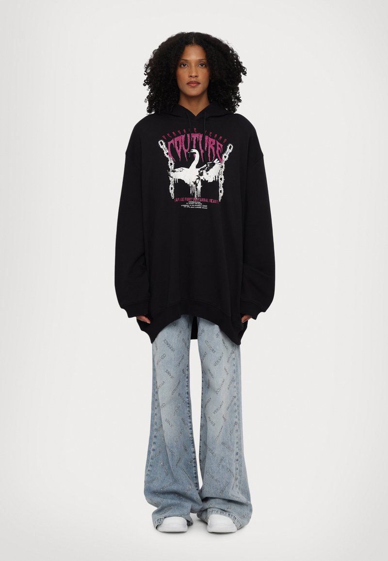 Black oversized hoodie with graphic design featuring white and pink text. Paired with light blue wide-leg jeans displaying branding.