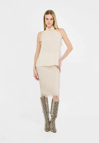 Haut beige sans manches avec détails volants, associé à une jupe crayon en dentelle ajustée et des bottes montantes à imprimé serpent, le tout dans des tons neutres.