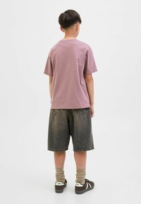Lila Baumwoll-T-Shirt, kurze Ärmel, lässige Passform. Kombiniert mit dunkelblauen Jeansshorts, kniehoch. Trägt hellbraune Socken und Sneakers.