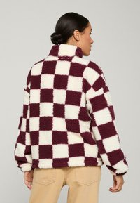Fleece bunda s vysokým límcem, která má burgundský a krémový kostkovaný vzor. Měkká textura a volný střih.