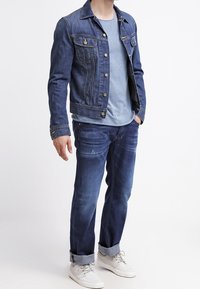 Giacca di jeans a lavaggio scuro, con chiusura a bottoni e tasche anteriori, abbinata a una t-shirt azzurro chiaro e jeans di jeans scuri, completata da sneakers bianche.