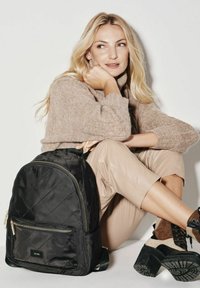 Czarny pikowany plecak z złotymi akcesoriami i przednią kieszenią na zamek. Modelka ma na sobie jasnobrązowy sweter i beżowe skórzane spodnie, siedząc przy białej ścianie.
