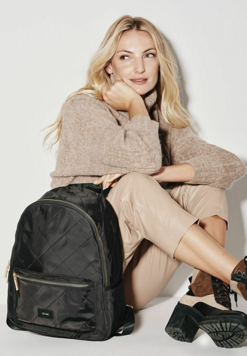 Czarny pikowany plecak z złotymi akcesoriami i przednią kieszenią na zamek. Modelka ma na sobie jasnobrązowy sweter i beżowe skórzane spodnie, siedząc przy białej ścianie.