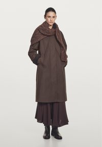 Brown waterproof coat with a loose fit, featuring large pockets and a high collar. Wrapped in a textured brown scarf. Black ankle boots.