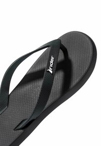 Chancla de goma negra con plantilla texturizada, que presenta una correa en forma de Y y el logo de la marca en blanco. Diseño suave y flexible.