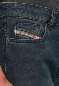 Detail kapsy tmavě modrých džínů ukazující přišitou štítek Diesel Industry Denim Division uvnitř.