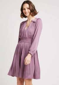 Robe violette à manches longues, taille smockée et jupe plissée. Présente des accents froncés au niveau du col et un tissu texturé.