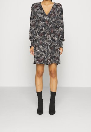 Femme portant une robe noire à motif floral avec col en V et manches longues, associée à des bottines chaussettes noires, debout devant un fond uni.