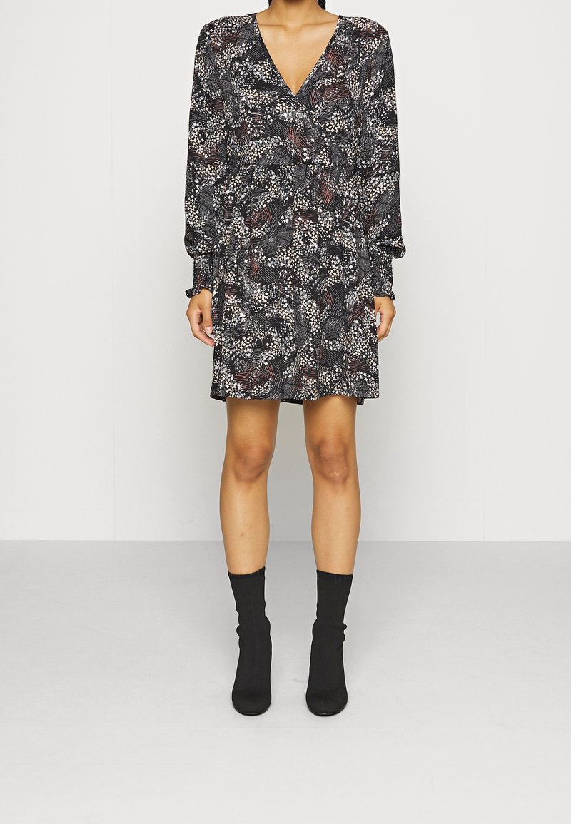 Femme portant une robe noire à motif floral avec col en V et manches longues, associée à des bottines chaussettes noires, debout devant un fond uni.