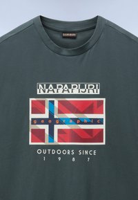 Tmavě zelené bavlněné tričko s grafickým potiskem, který zobrazuje vrstvené vlajky, slovo "NAPAPIJRI" a "OUTDOORS SINCE 1987" v bílé barvě.