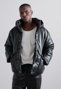 Čierna puffa bunda so lesklou textúrou, oversized kapucňou a viditeľnými cvokmi, skombinovaná s bielou priliehavou košeľou pod ňou.