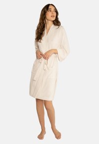 Accappatoio beige chiaro realizzato in materiale morbido, con maniche lunghe, una cintura in vita e due tasche frontali.