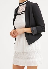 Czarna niezbyt długa marynarka uszyta z gładkiego materiału, połączona z białą sukienką z koronkowymi detalami i wyższym kołnierzykiem. Krótkie rękawy, warstwowy krój.