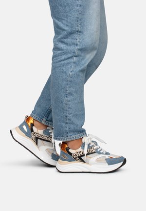 Person iført lyseblå jeans og hvide sneakers med orange, blå og dyreprintdetaljer, stående med den ene fod let løftet.