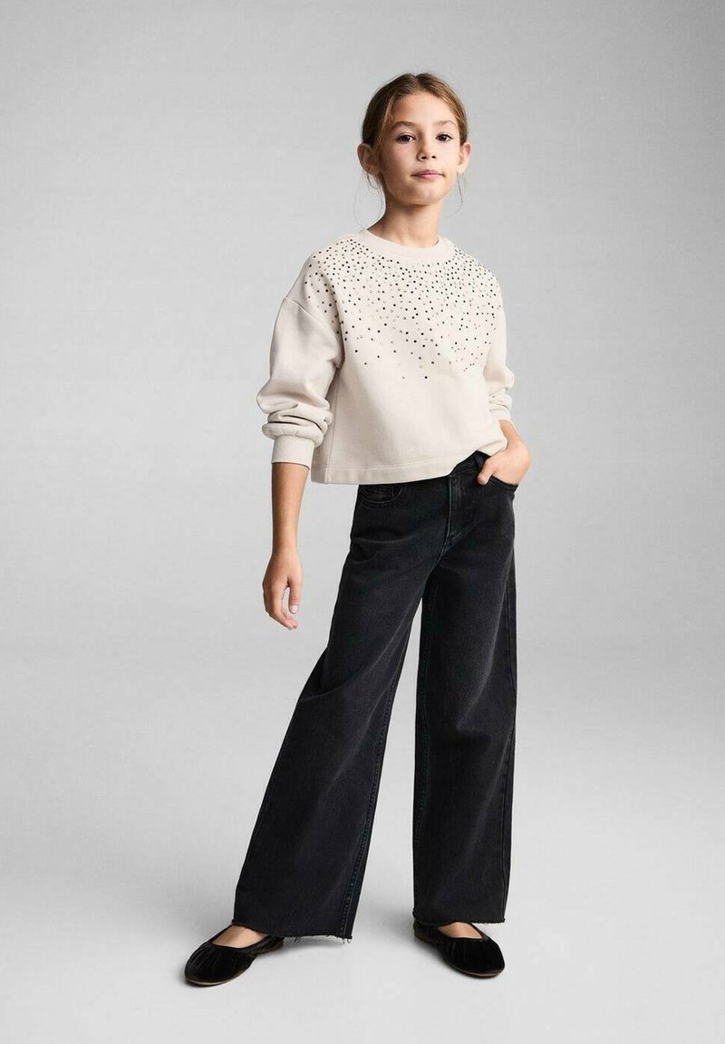 Felpa beige chiaro con decorazioni in rivetti, abbinata a jeans neri a gamba larga. Il modello è in posa con una mano nella tasca, le scarpe sono ballerine nere.