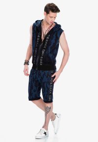 Marineblaues ärmelloses Hoodie und passende Shorts mit einem strukturierten Muster. Enthält Logos in kontrastierenden Farben. Weiße Turnschuhe mit Akzenten.