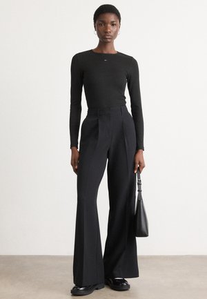 Black long-sleeve top paired with high-waisted, wide-leg black trousers. Model holds a small black handbag. Smooth fabric textures.