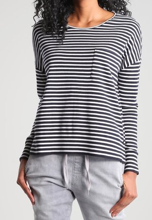 Femme portant un tee-shirt à manches longues à rayures horizontales noires et blanches avec une petite poche sur la poitrine et un pantalon gris clair à cordon de serrage.