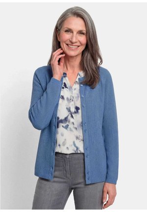 Vrouw van middelbare leeftijd die lacht, draagt een blauw vest met knopen over een witte blouse met patroon en grijze broek, met lang grijs haar.
