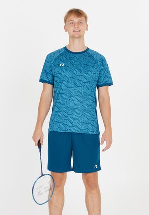 Ung mand står i blå mønstret sportstøj med et badmintonracket mod en ensfarvet hvid baggrund.