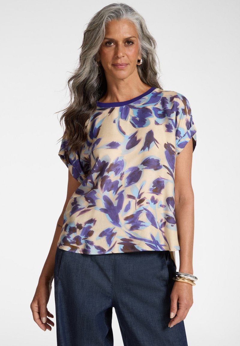 Volwassen vrouw met lang grijs haar, gekleed in een beige top met korte mouwen en paarse bloemenprint, en donkerblauwe broek, voorzien van armbanden.
