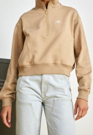 Osoba v béžové mikině s půlzipem a světle modrých džínách s vysokým pasem, stojící s rukama volně podél těla.