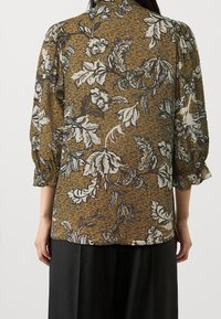 Bruine bloemenblouse met lange mouwen, voorzien van witte en zwarte blaadjespatronen en een gestructureerde achtergrond. Gedrapeerde silhoët, lichtgewicht stof.