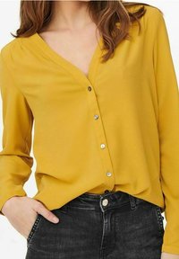 Blouse jaune à manches longues avec un col en V et des boutons sur le devant, confectionnée dans un tissu lisse. Portée avec un jean noir, offrant une coupe décontractée.