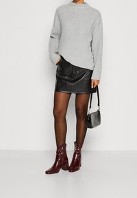 Žena v šedém žebrovaném svetru, černé kožené mini sukni, průhledných černých punčochách, bordo kotníkových botách a držící černou kabelku.